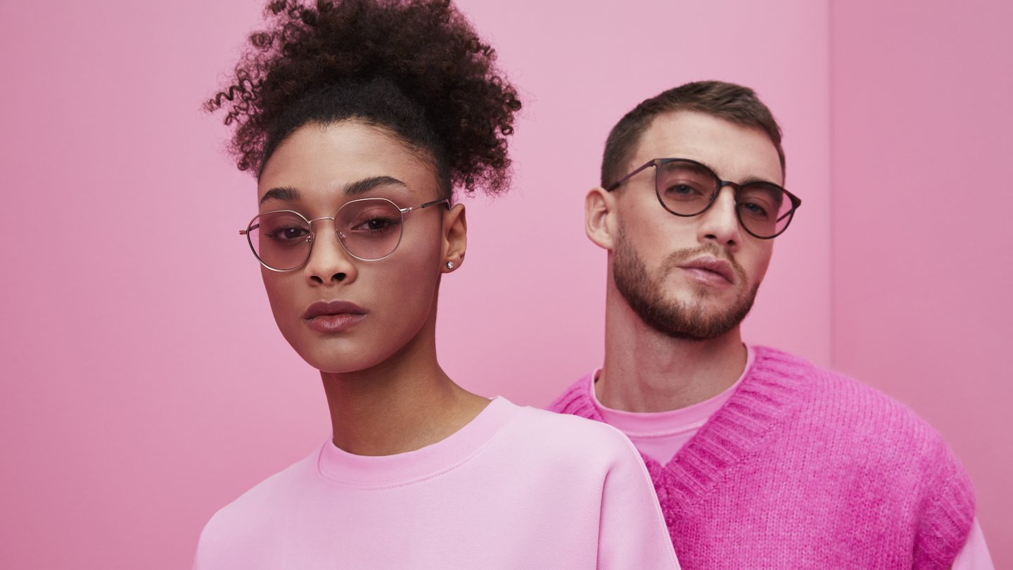 Das Foto zeigt zwei Personen, die eine Brille mit Filtergläsern tragen, vor einem rosafarbenen Hintergrund.