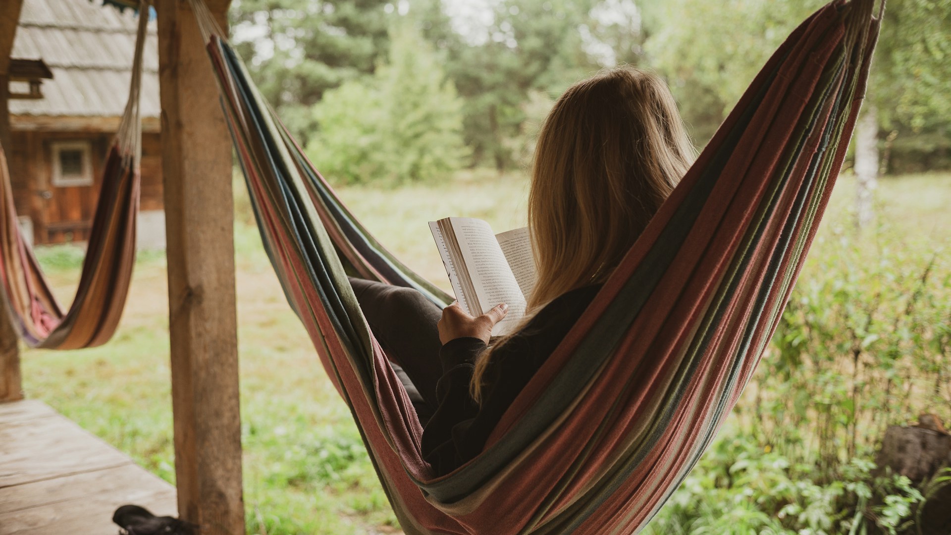 Eine Frau liegt in einer Hängematte, die an einer Holzterrasse zwischen zwei Balken befestigt ist. Sie liest ein Buch und ist von hinten fotografiert, sodass ihr Gesicht nicht sichtbar ist.