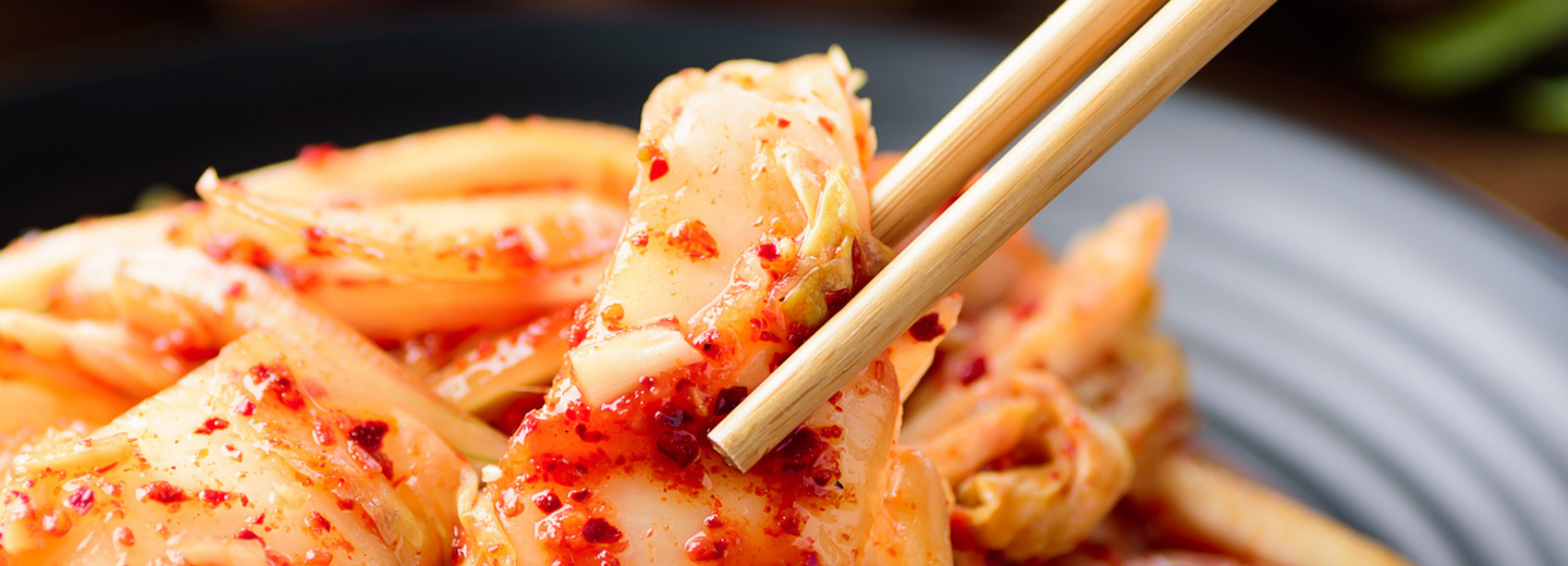 Ein Teller, auf dem Kimchi liegt und Stäbchen, die ein Stück Kimchi halten. 