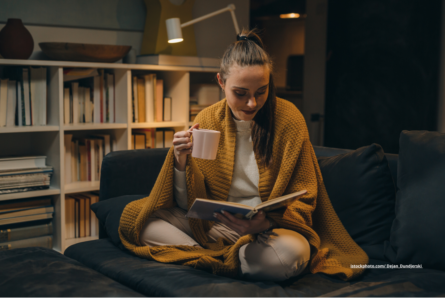 Eine Frau, die auf dem Sofa sitzt und liest. In einer Hand hält sie ein Buch, in der anderen eine Tasse. 