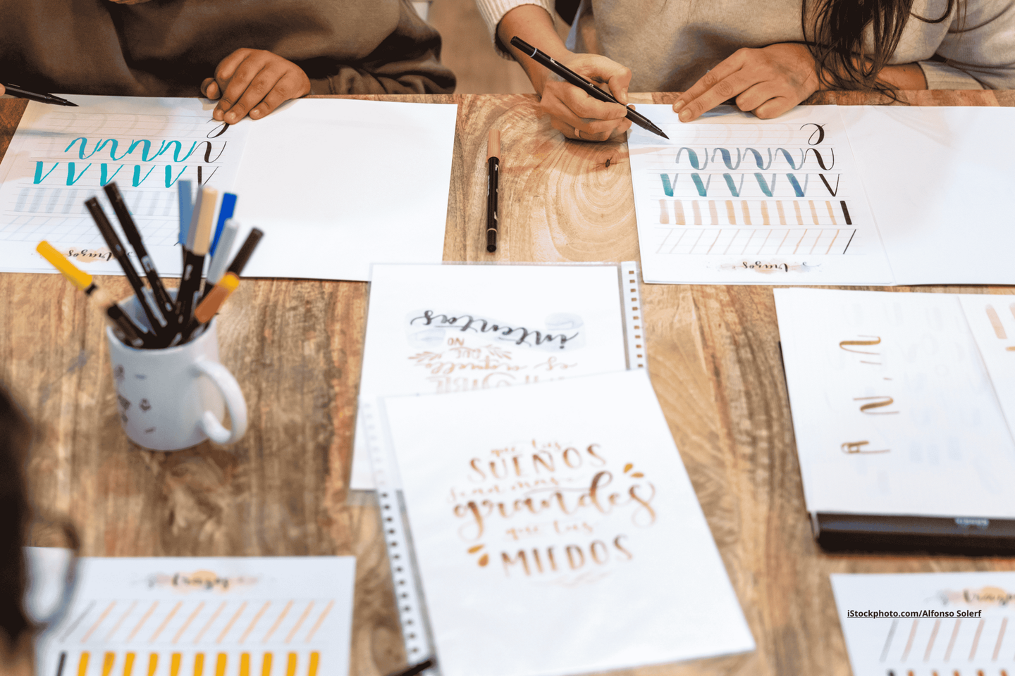 Ein Tisch auf dem Blätter mit Kalligrafie Schriftbögen liegen und eine mit Stiften gefüllte Tasse stehen. Es sind zwei Paar Hände, die Kalligrafie üben zu sehen. 