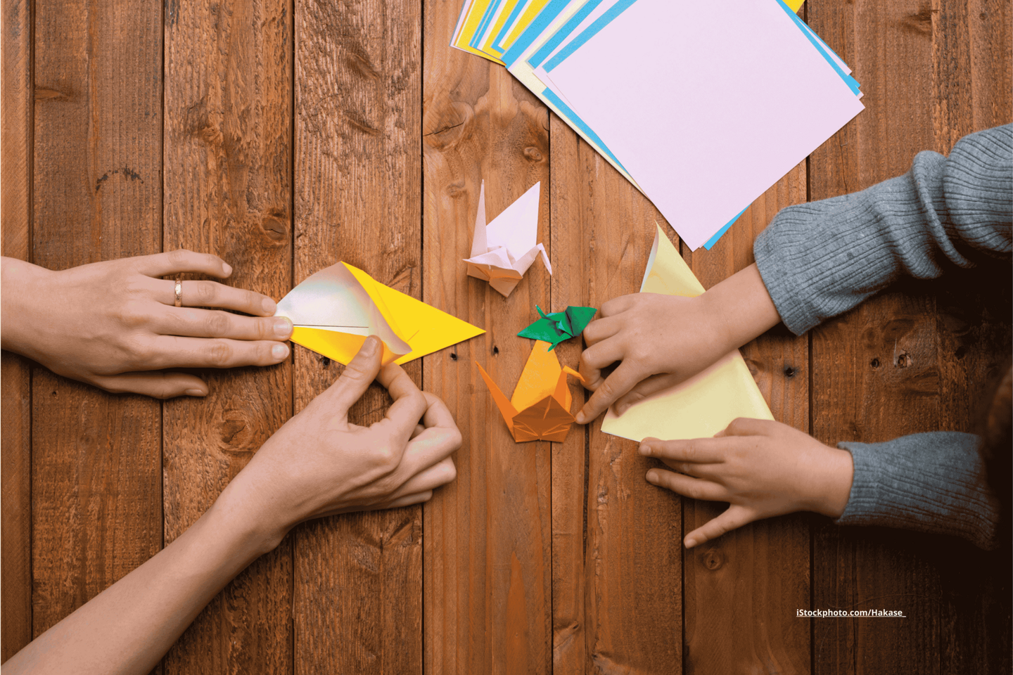 Zwei Paar Hände, von einer Erwachsenen Person und einem Kind, die buntes Origami Papier auf einem Holztisch falten. Zwischen ihnen stehen fertig gefaltete Kraniche. 