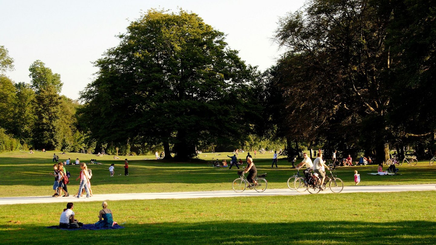 Menschen genießen einen sonnigen Tag im grünen Park, mit Spaziergängern und Radfahrern auf dem Weg, Kinder, die spielen, und Gruppen, die auf dem Rasen entspannen.