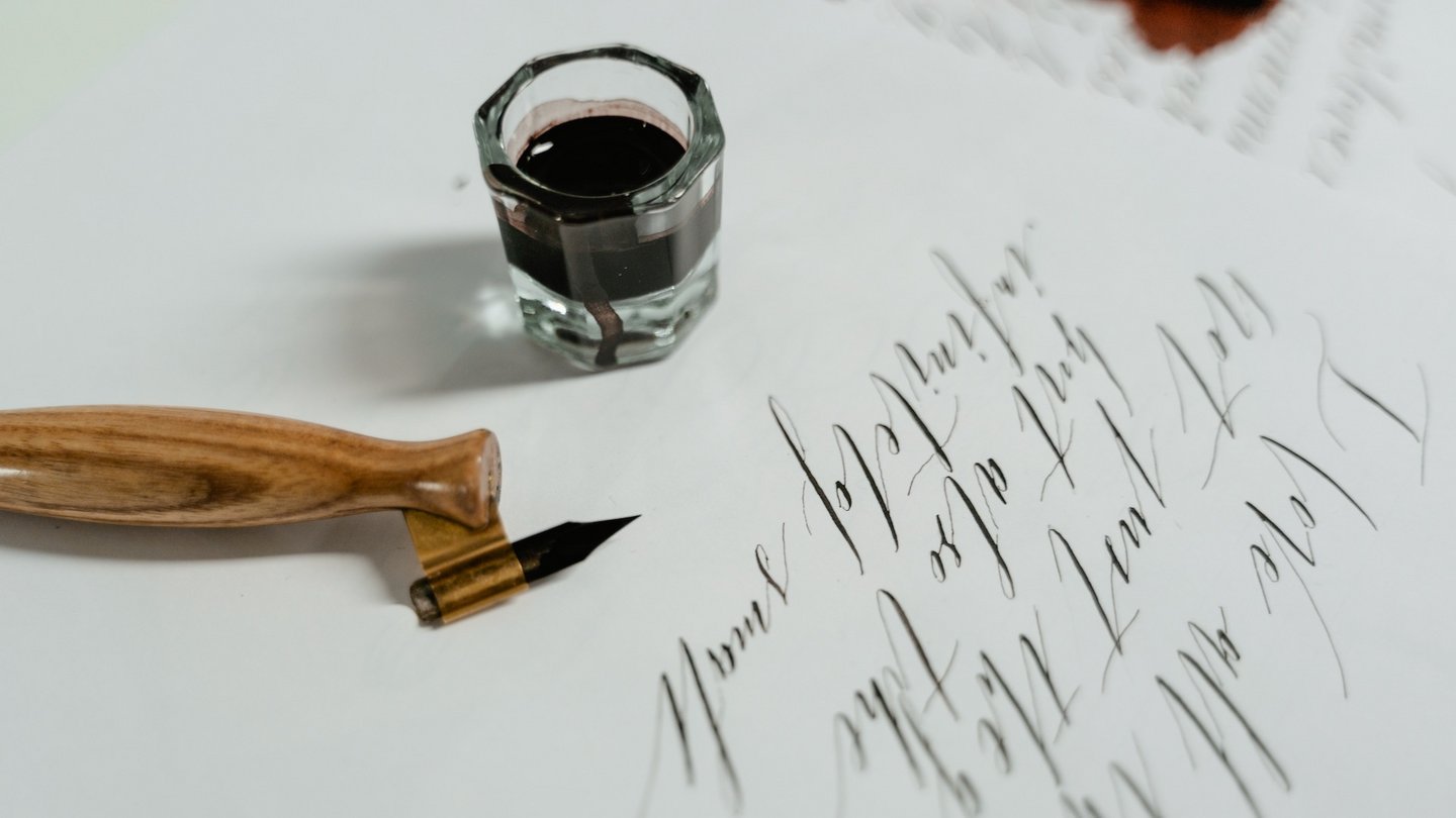Das Bild zeigt einen Federhalter und ein Tintenfässchen auf einem Blatt Papier.