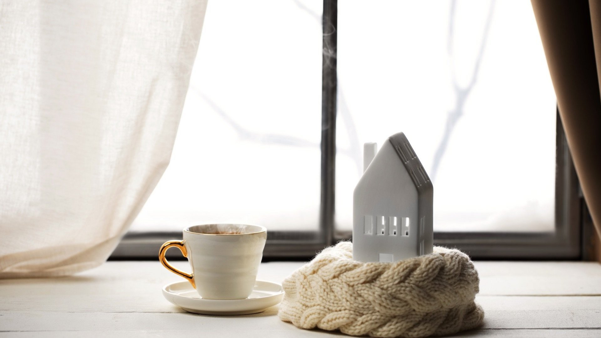 Das Foto zeigt eine dampfende Teetasse und ein Keramik-Haus vor einem Fenster.