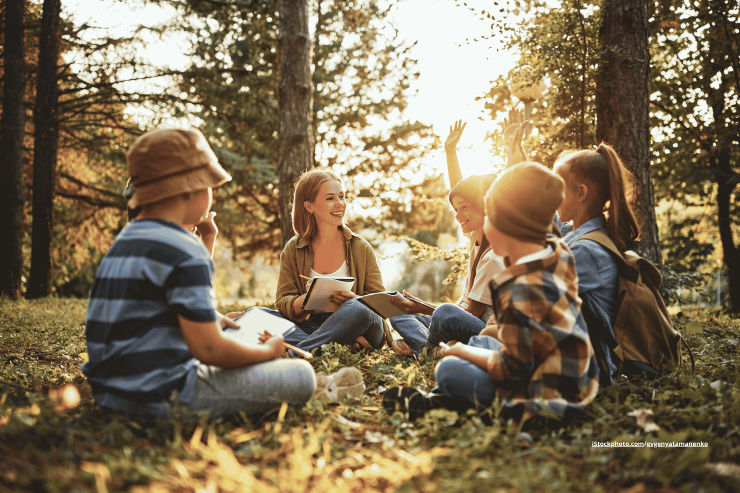 Eine Frau und fünf Kinder, die im Wald auf dem Boden sitzen und Hefte in den Händen halten. 