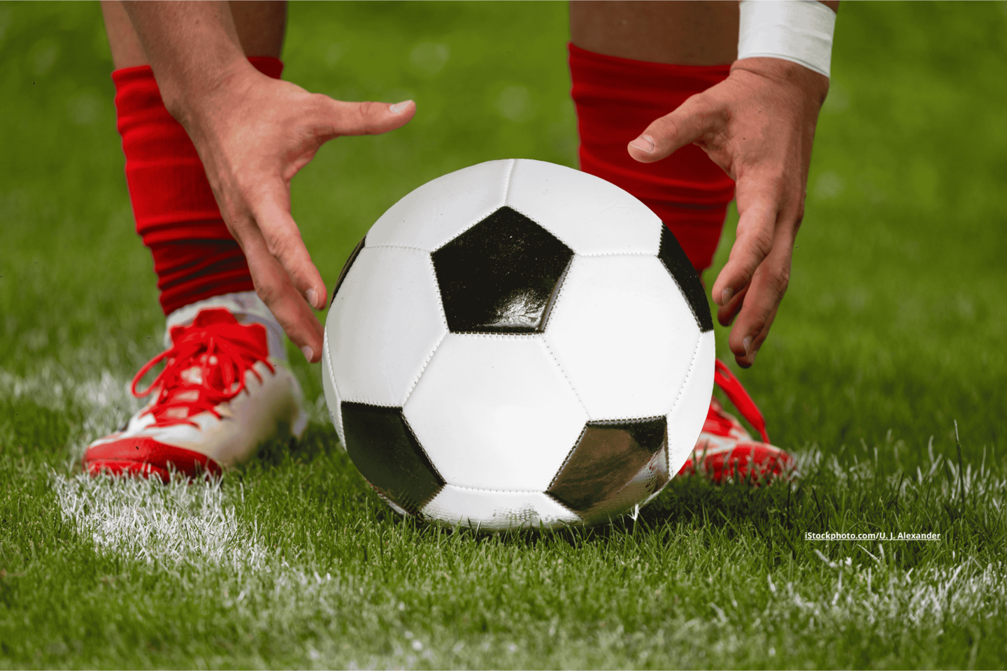 Ein Fußball, der auf dem Rasen liegt, hinter ihm sind die Füße und Hände eines Fußball-Spielers zu sehen.