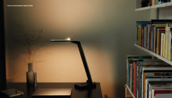 Das Foto zeigt die Tischleuchte elumentis® LED von Eschenbach Optik auf einem Schreibtisch stehend und warm leuchtend. 
