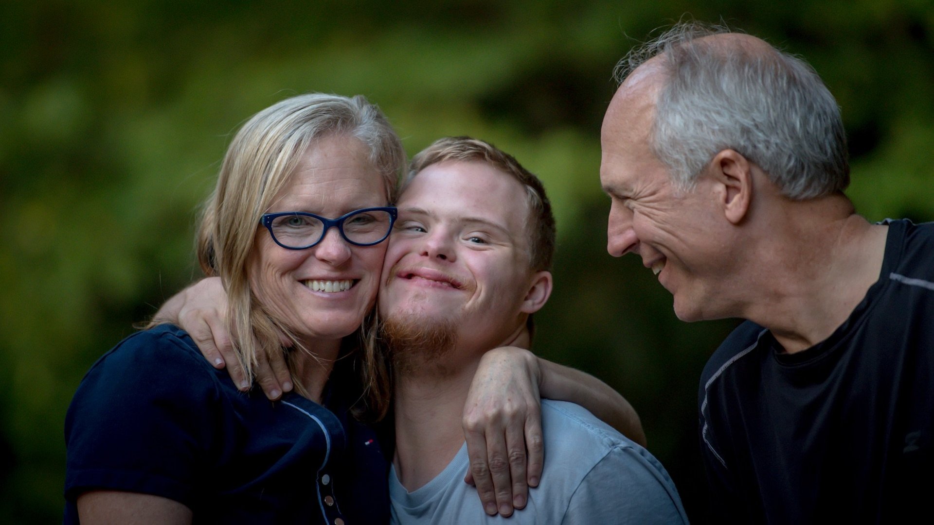Da Bild zeigt eine lachende Familie, in der Mitte eine Person mit Down Syndrom.