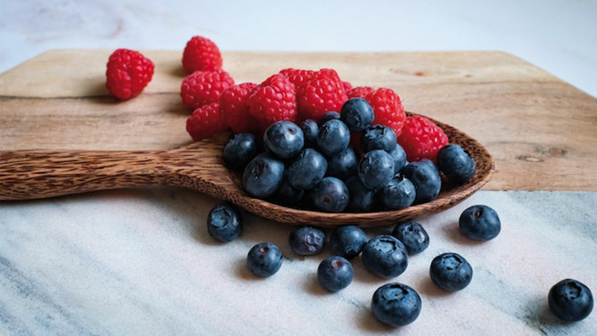 Blaubeeren und Himbeeren in einem Holzlöffeln vor einem Holzbrett