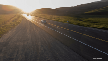 Das Foto zeigt eine Landstraße, auf der zwei Autos bei tief stehender Sonne fahren.