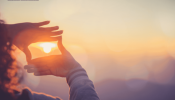 Zu sehen sind die Hände einer Frau, die einen Rahmen um die aufgehende Sonne formen. Der Himmel ist orange. 