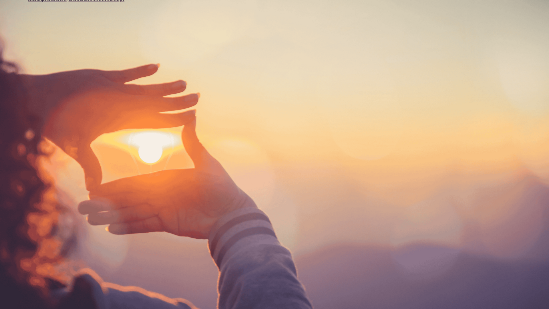 Zu sehen sind die Hände einer Frau, die einen Rahmen um die aufgehende Sonne formen. Der Himmel ist orange.