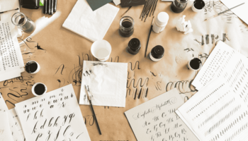 Eine Arbeitsfläche, auf der diverse Kalligrafie-Utensilien liegen, u.a.: Tintenfässer, ein Stift, mehrere Papierbögen. 