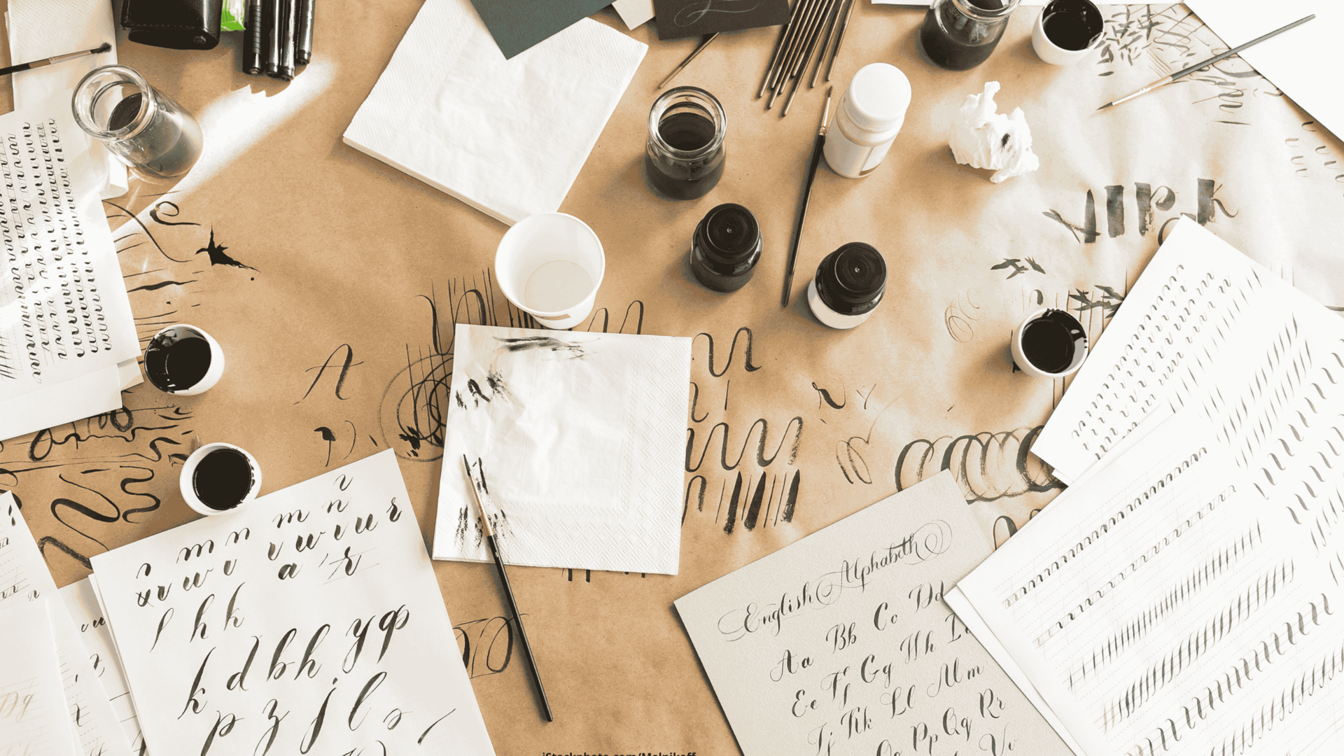 Eine Arbeitsfläche, auf der diverse Kalligrafie-Utensilien liegen, u.a.: Tintenfässer, ein Stift, mehrere Papierbögen. 