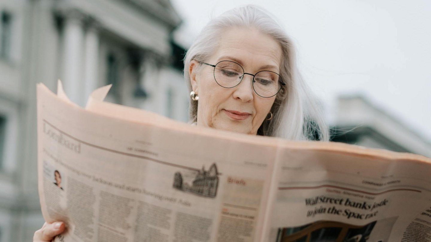 Eine ältere Dame mit grauem Haar und runder Brille vertieft in die Lektüre einer Zeitung, mit einem fokussierten und nachdenklichen Gesichtsausdruck.