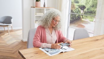 Eine Frau mit weißem lockigen Haar liest konzentriert ein Magazin mit einer elektronischen Lupe von ESCHENBACH OPTIK GmbH an einem Holztisch in einem hellen Raum mit Blick auf den Garten.