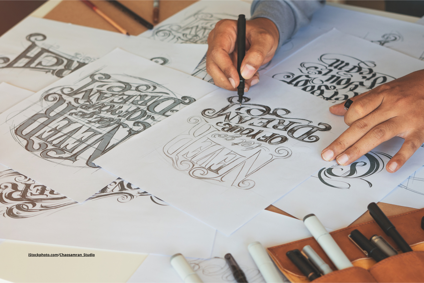 Ein Tisch, auf dem Blätter liegen, auf welchen Handlettering zu sehen ist. Ein Paar Hände, das einen Stift hält und eine Handlettering Übung macht.