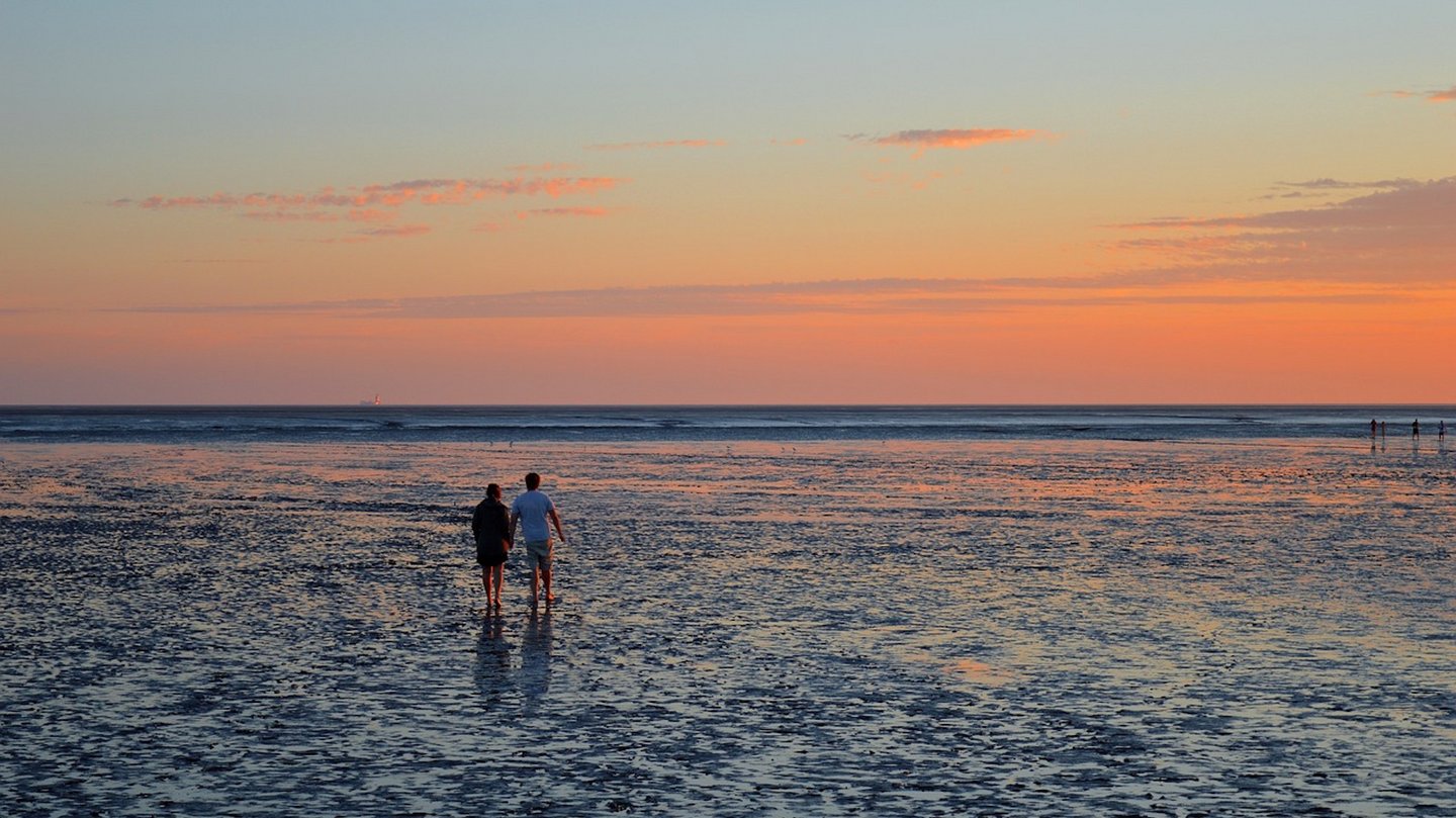 Das Bild zeigt ein Paar während eines Sonnenuntergangs am Wattenmeer spazieren.