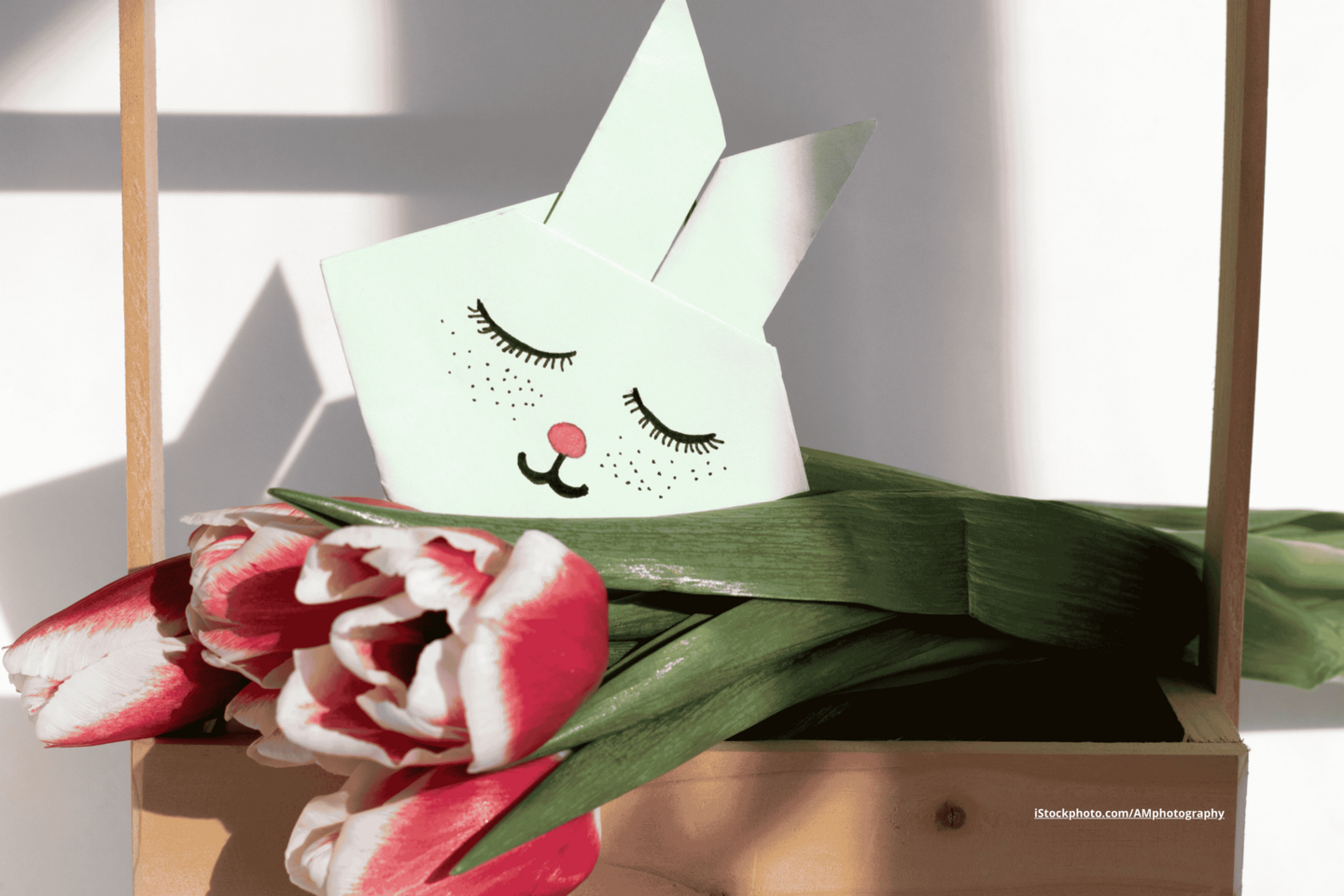 Ein pastellgrüner Origami Hasenkopf, platziert auf einem liegendem Strauß rot-weißer Tulpen. 