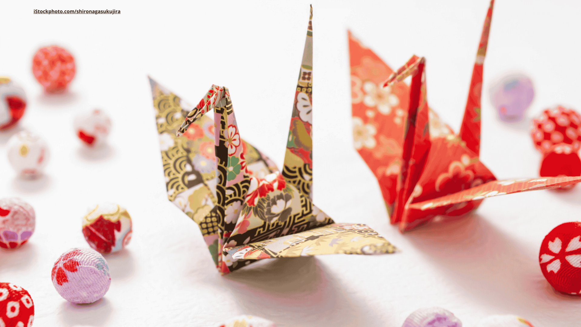 Zwei Origami Kraniche aus bunt gemustertem Papier, stehend auf ebenfalls gemustertem Origami Papier.