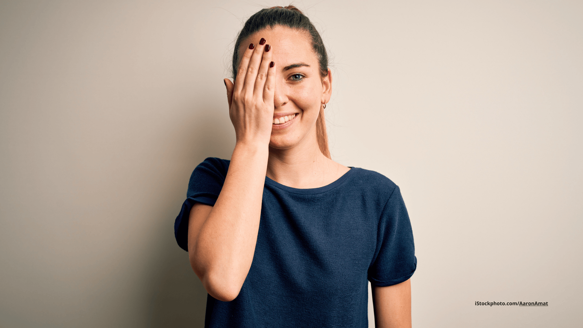 Das Foto zeigt eine Frau vor weißem Hintergrund, die sich ein Auge mit der Hand zuhält und dabei lächelt.